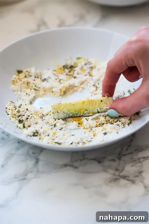 A close-up shot of a hand dipping a slice of zucchini into a bowl of dry seasoning mix, demonstrating the coating process for the fries.