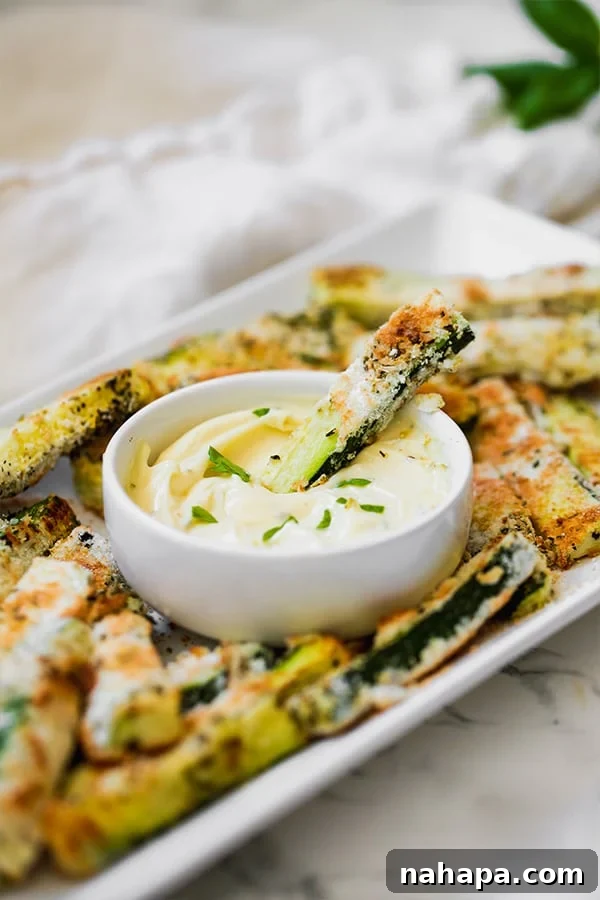 A white serving dish of golden baked zucchini fries, with a small white bowl of creamy dipping sauce in the center, and one fry being dipped into the sauce.