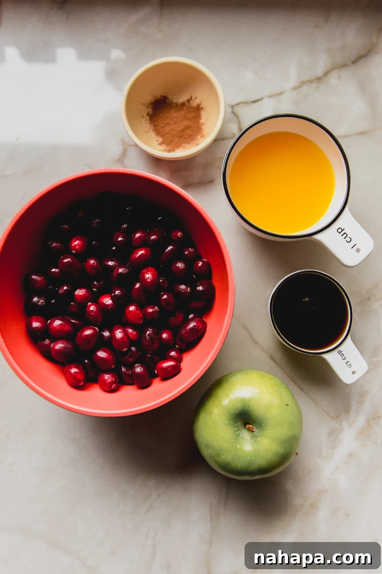 Spiced Apple Cranberry Compote 3 All the fresh ingredients for apple cranberry sauce laid out in separate small bowls: cranberries, diced apple, orange juice, maple syrup, cinnamon, and orange zest.