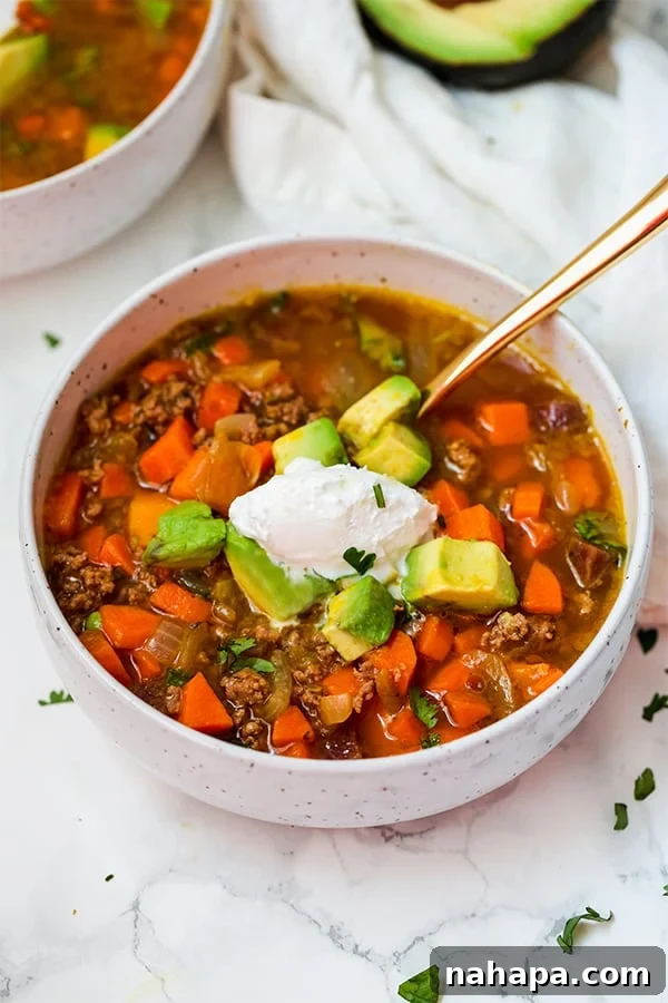 A white bowl filled with savory Paleo & AIP pumpkin chili, generously topped with creamy coconut cream and perfectly sliced avocado.