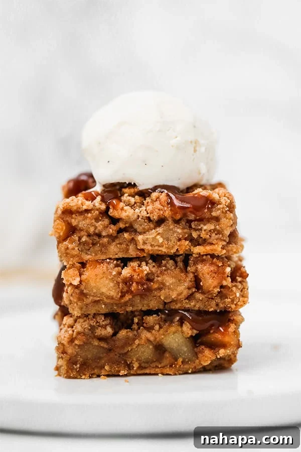 A stack of three apple pie bars topped with a scoop of vanilla ice cream and a drizzle of caramel sauce.