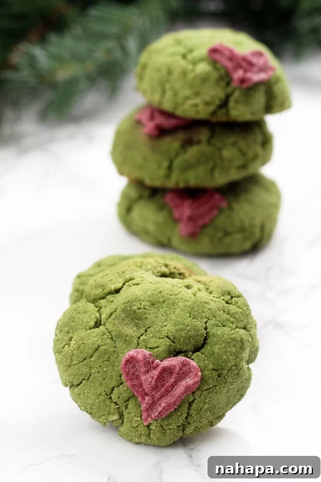 A pile of green Paleo Grinch Cookies, some with a red heart decoration, on a festive background.