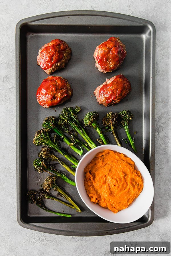 A sheet pan with roasted broccolini, mini meatloaves, and a white bowl of mashed sweet potatoes.