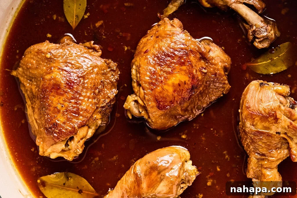 Chicken simmering in a rich, dark sauce with bay leaves in a pan.