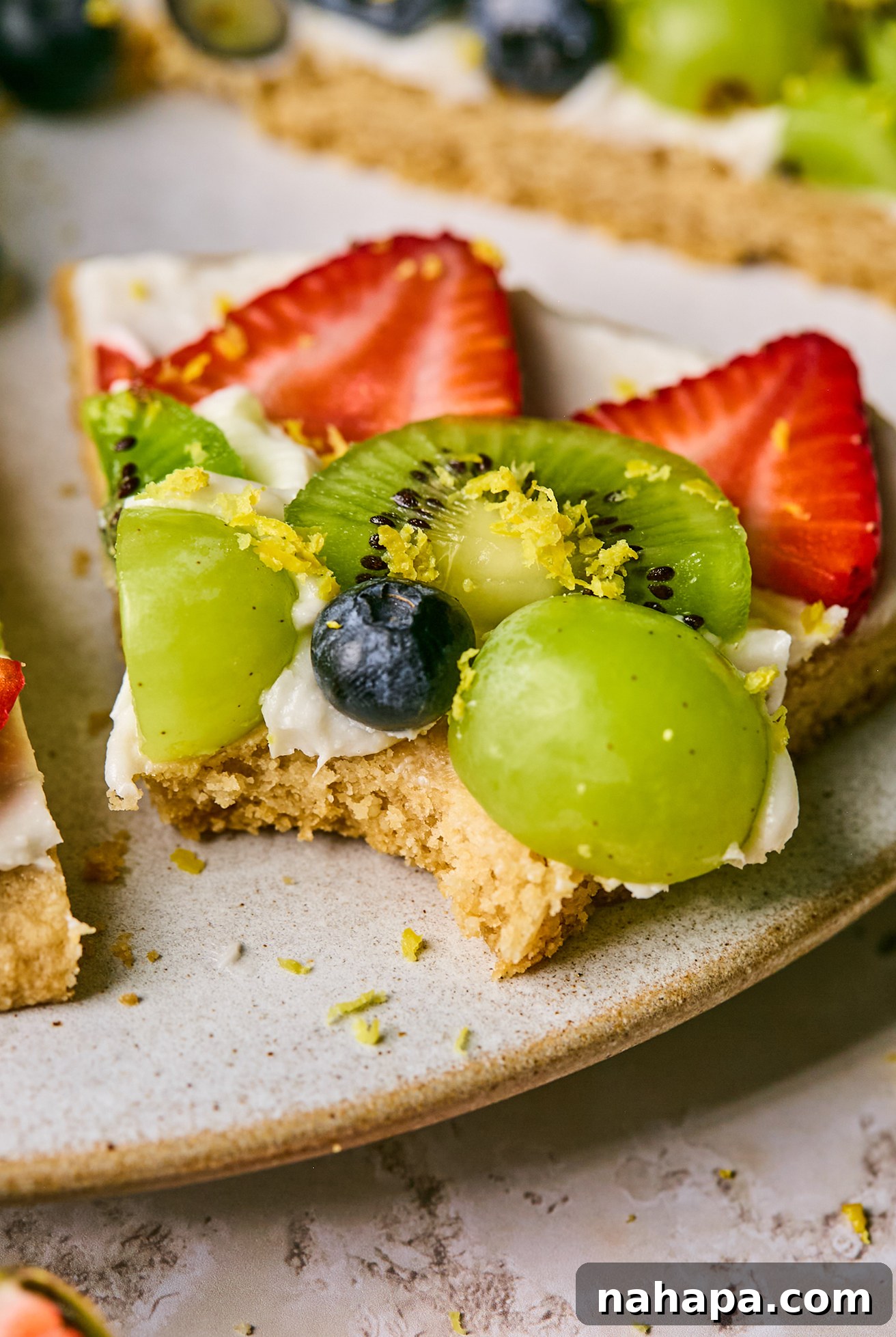 A slice of sugar cookie fruit pizza with a bite taken out of it, showcasing the cookie, frosting, and fruit layers.