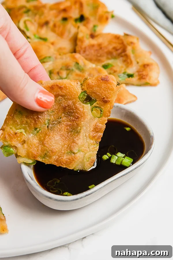 A hand dipping a quarter of a crispy gluten-free scallion pancake into a small bowl of coconut aminos dipping sauce, highlighting the texture and serving suggestion.