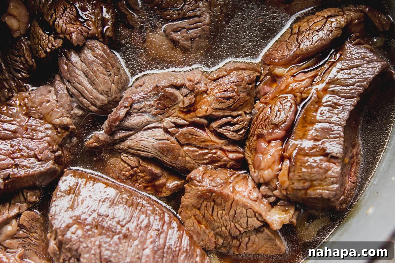 Chuck roast pieces cooked with sliced onion in liquid in a Dutch oven, surrounded by the savory sauce ingredients.