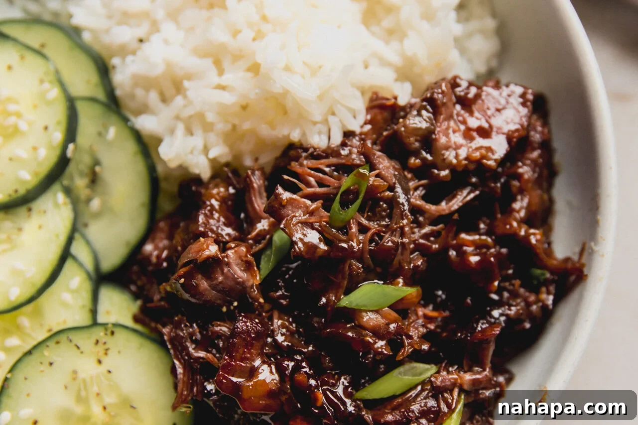 A white bowl with ginger chuck roast next to white rice and cucumber slices, ready to be served.