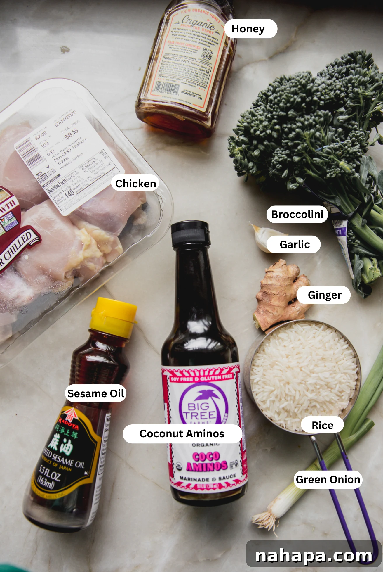 A flat lay photograph showcasing all the fresh ingredients for brothy ginger chicken and rice, including raw chicken thighs, fresh ginger, garlic, green onions, broccoli, carrots, bone broth, coconut aminos, honey, and lime.