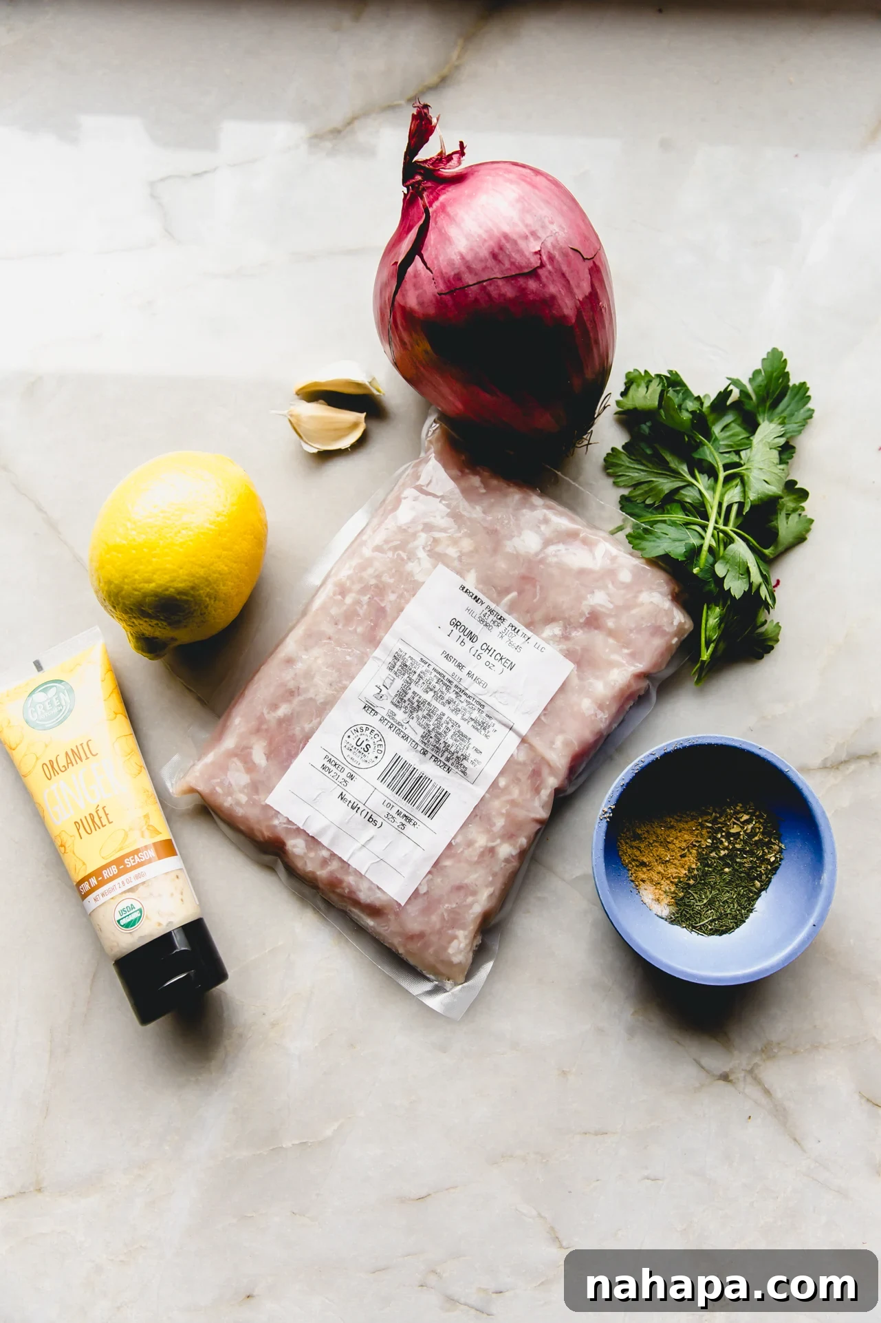 Sheet pan kebab ingredients laid out on a marble counter top, including ground chicken, spices, red onion, and lemon.