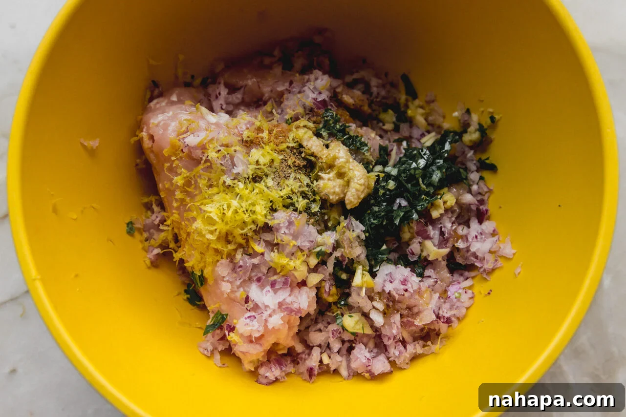 A yellow mixing bowl with ground chicken, seasonings, lemon zest, olive oil, and grated red onion before being mixed together.