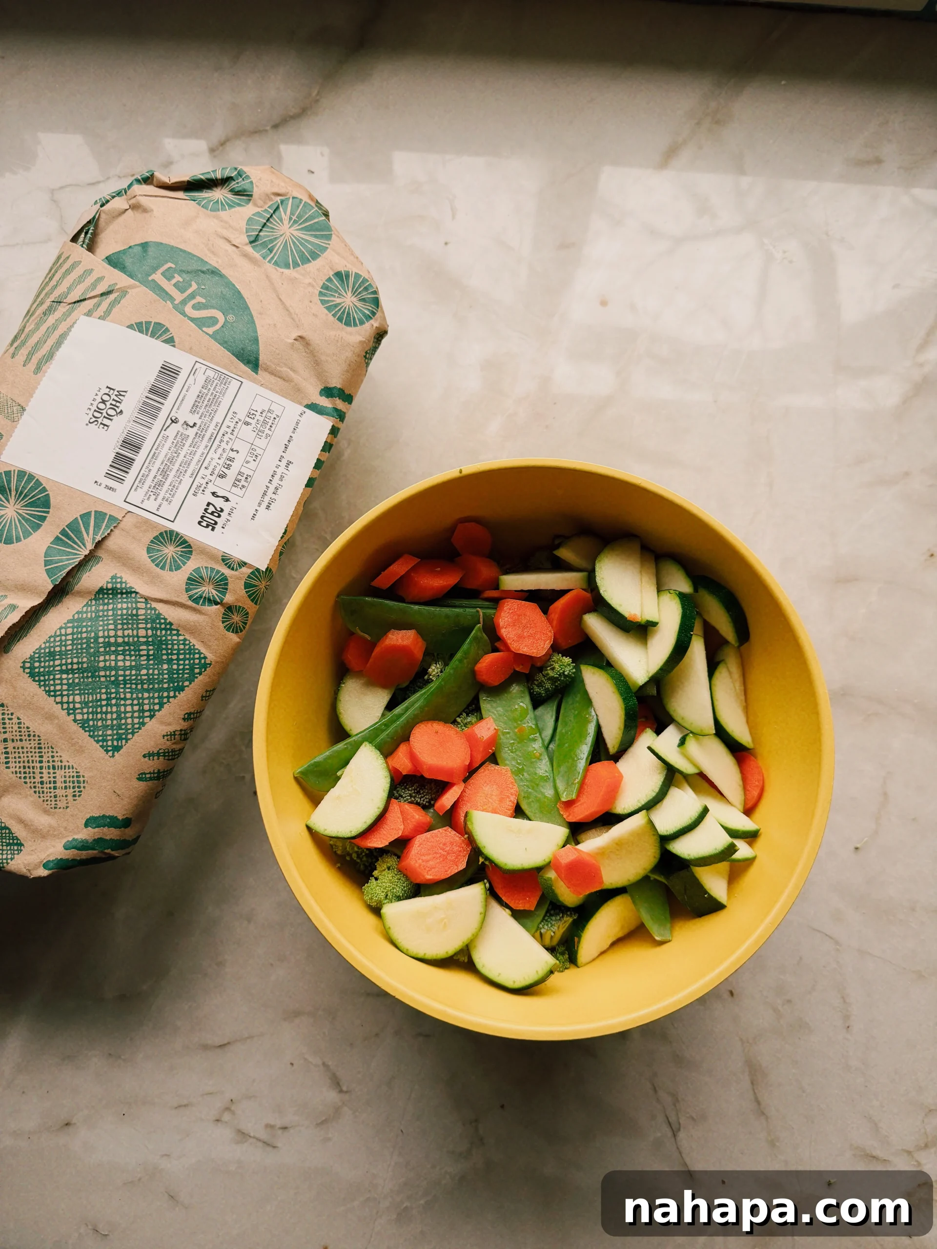 A flat lay photograph of all the raw ingredients neatly arranged on a kitchen counter, ready for the beef noodle stir fry. Ingredients include raw flank steak, a variety of fresh vegetables like snow peas, broccoli florets, sliced carrots, and chopped zucchini, gluten-free spaghetti, and bottles of coconut aminos and avocado oil.