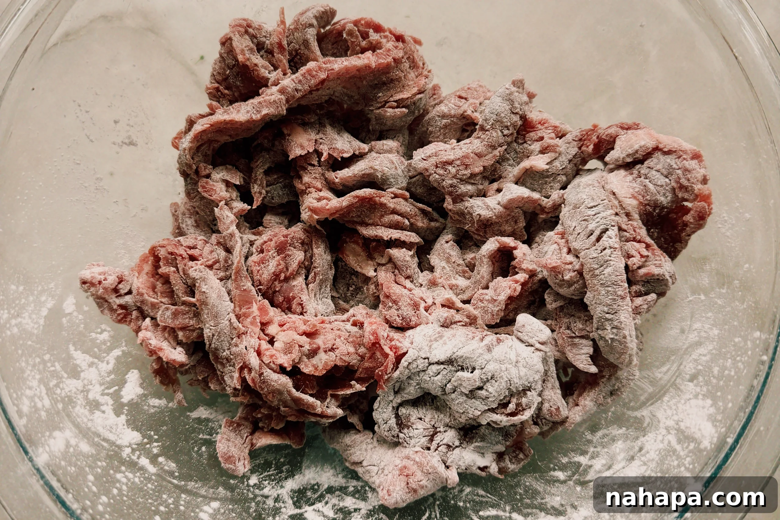 Thinly sliced flank steak in a clear glass bowl, generously coated with a light dusting of arrowroot starch, salt, and pepper, ready for cooking.