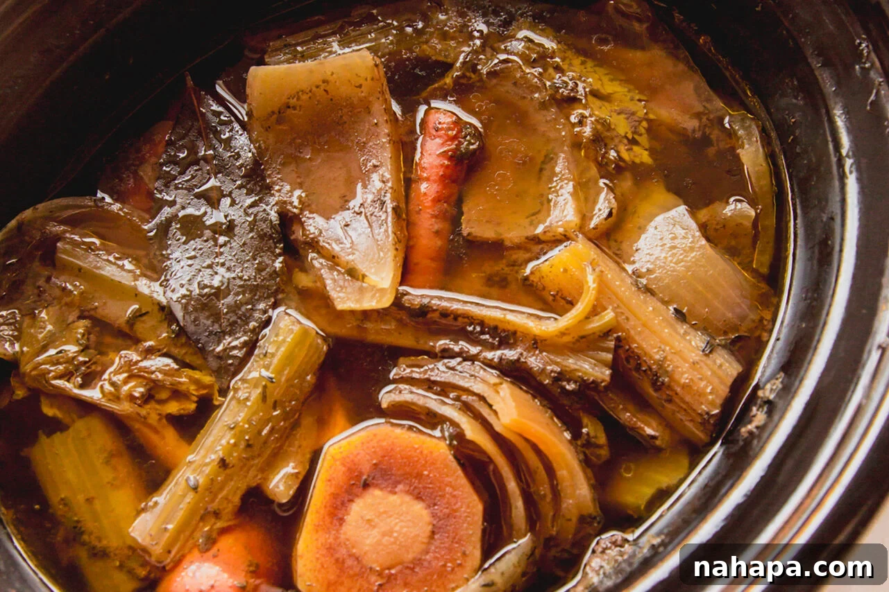 Beef bone broth simmering in a slow cooker, filled with roasted bones and an assortment of vegetable scraps.