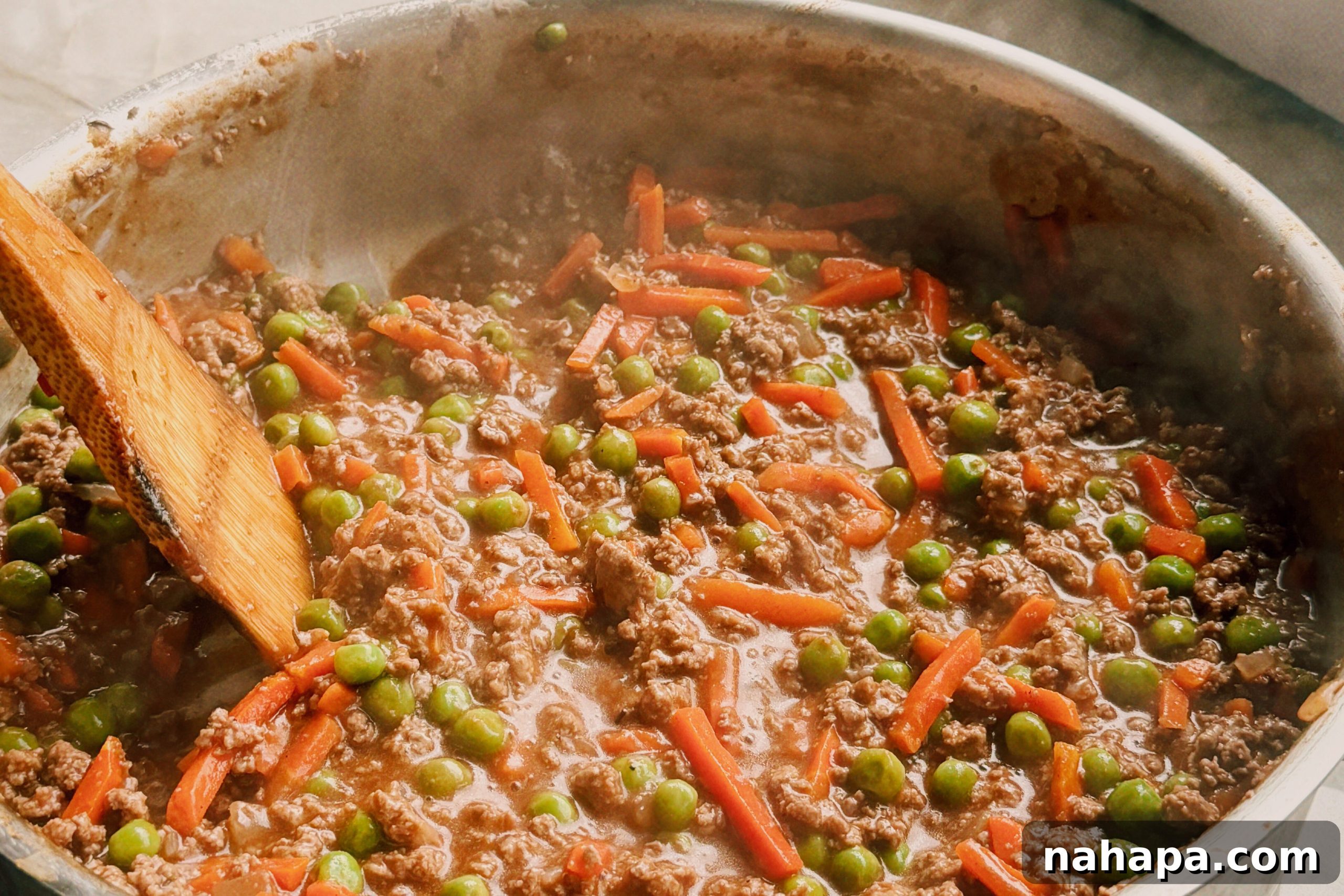 A saute pan with ground beef, peas and carrots, and a wooden spoon stirring it.