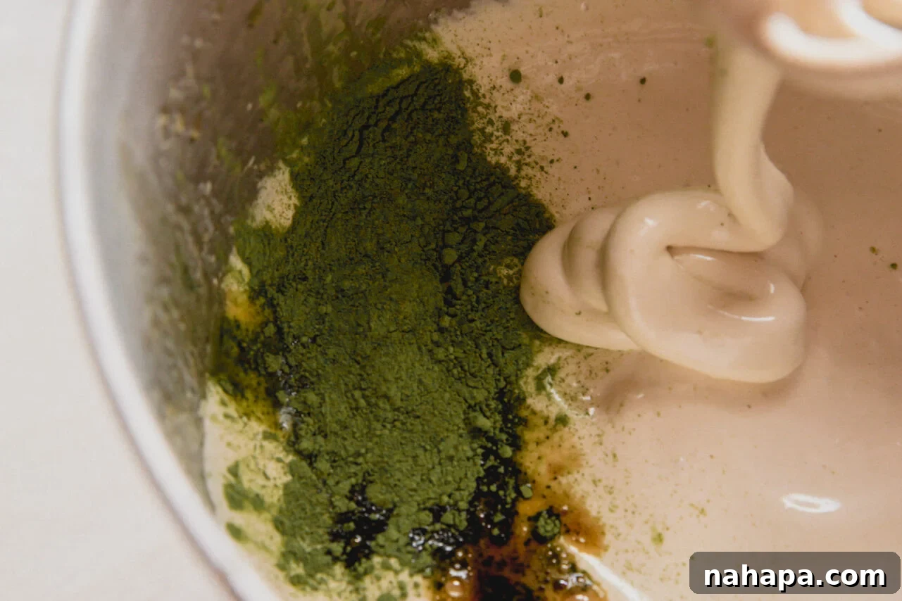 A stand mixer with the honey, water, and gelatin mixture after whipping, having vanilla and matcha powder added in, showcasing the vibrant green hue.