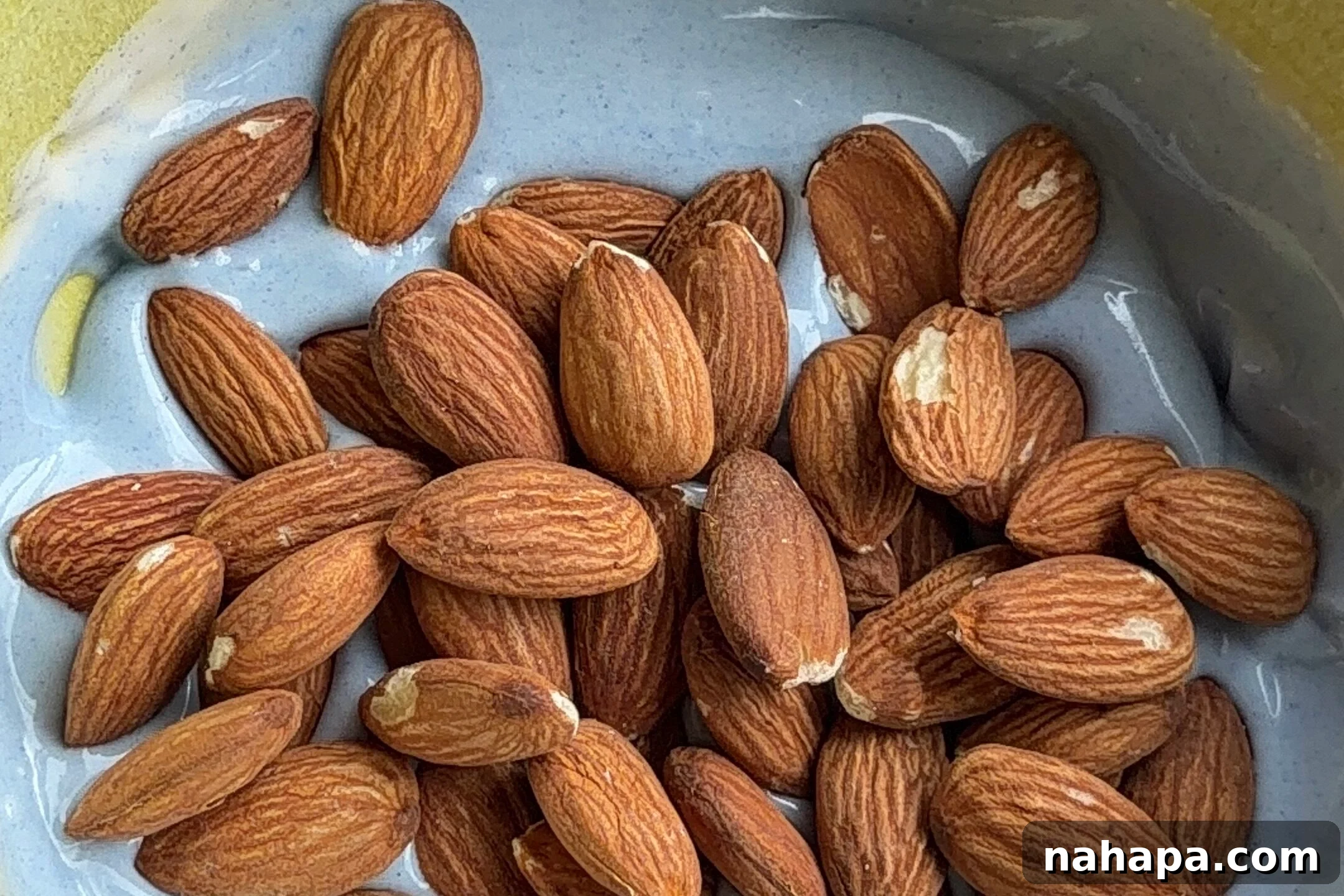 Raw almonds being added to a yellow bowl containing a creamy, melted dairy-free white chocolate and blue spirulina mixture, ready for coating.