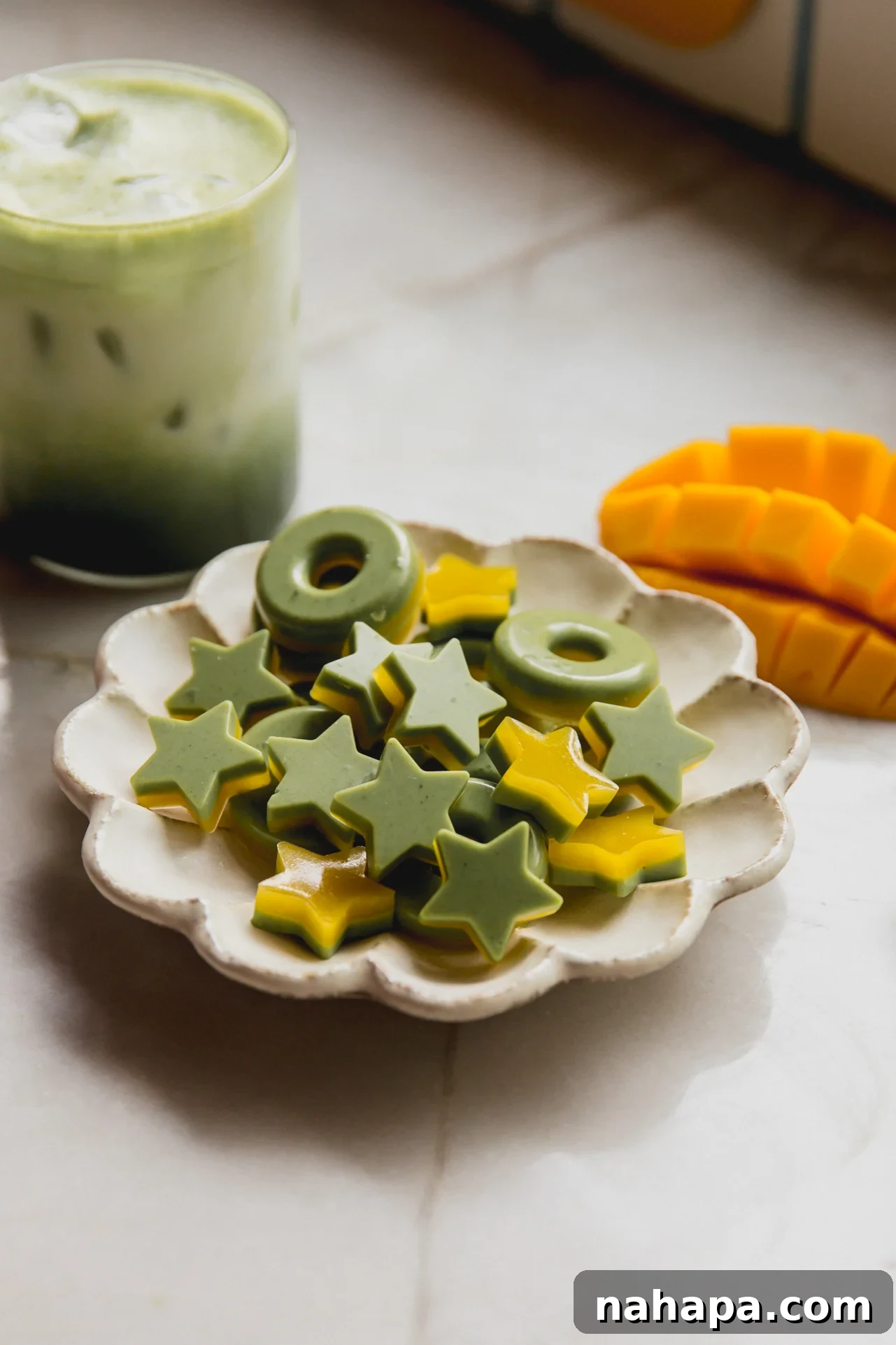 Star and ring shaped two tone mango and matcha gummies on a plate with a sliced mango and matcha latte in the background.