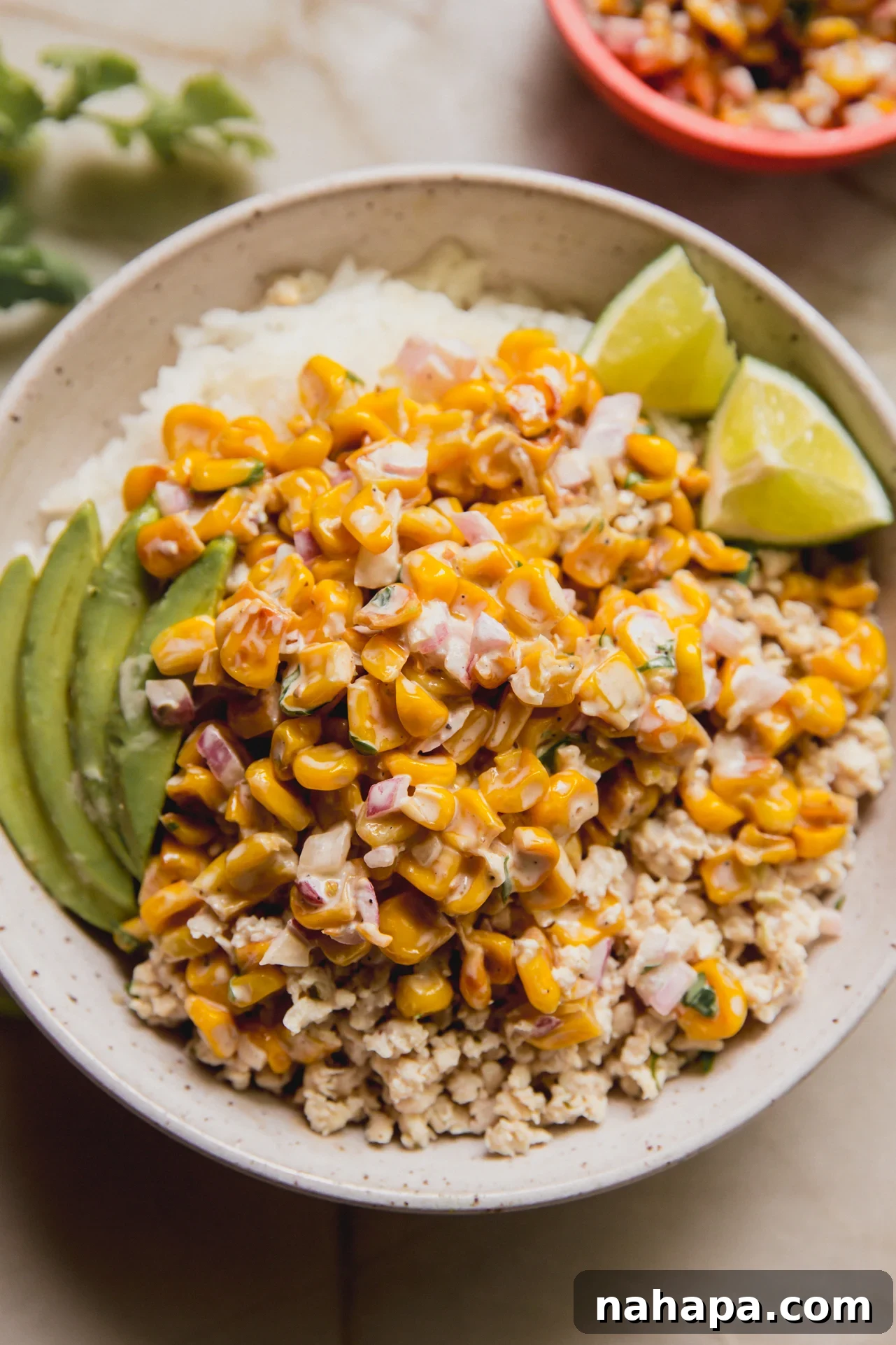 A vibrant bowl of rice and seasoned ground chicken, generously topped with creamy street corn, sliced avocado, and a fresh lime wedge. It's a colorful and inviting meal.