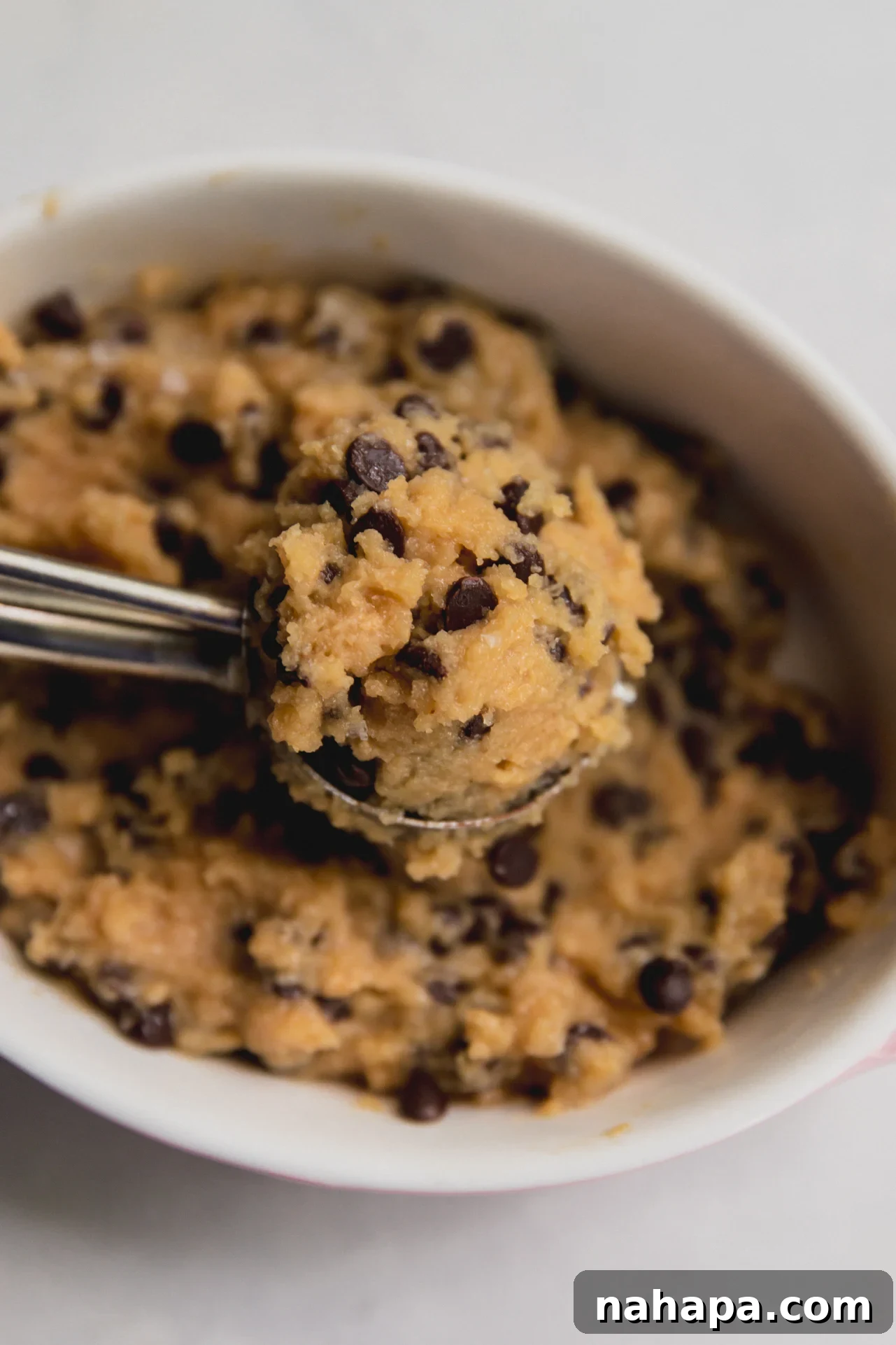 A white bowl of protein cookie dough with a metal scoop in it.