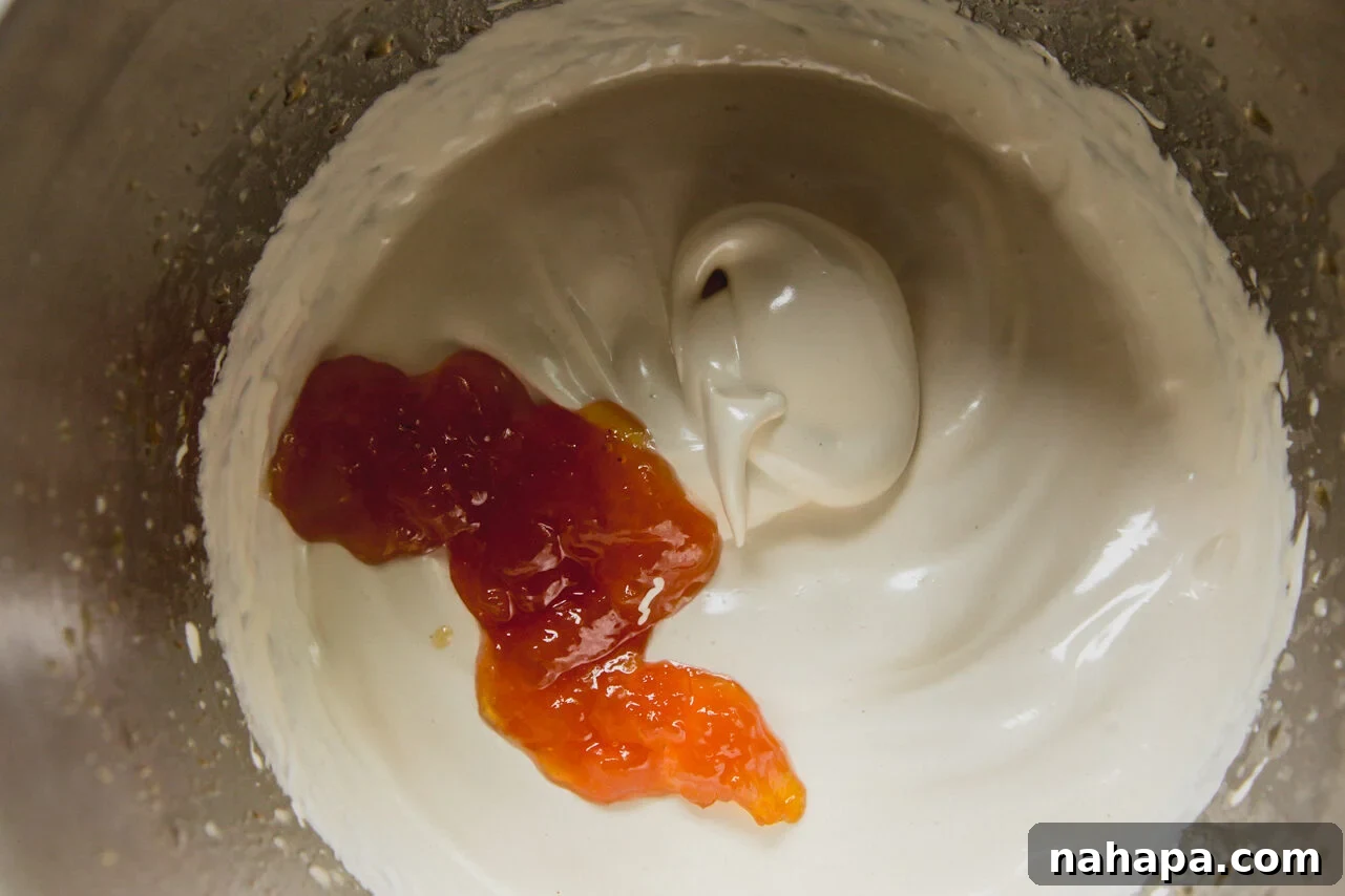 The stainless steel bowl of a stand mixer with marshmallow whip and peach jam being added to it.