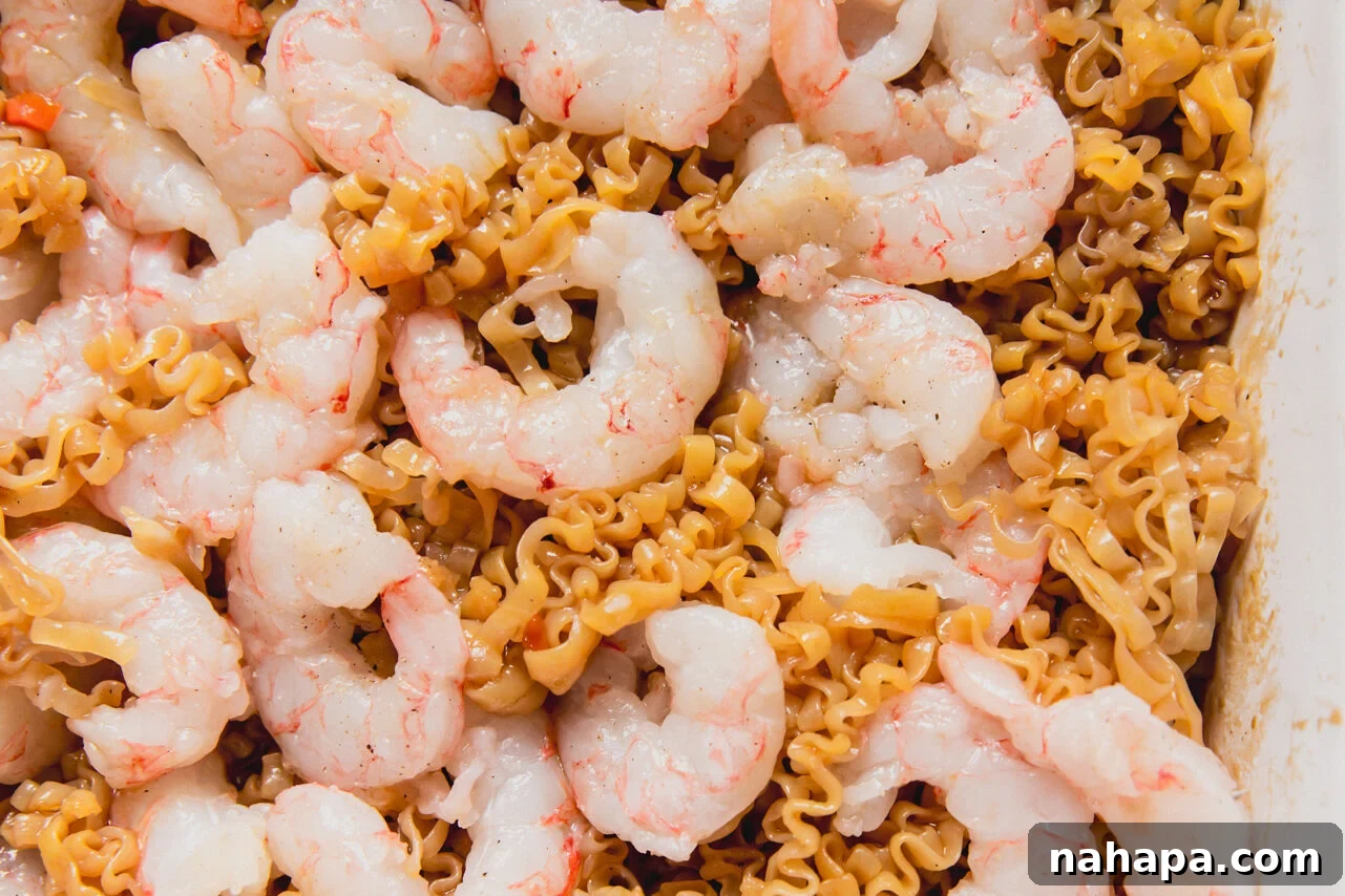A white casserole dish with raw shrimp on top of a ramen and vegetable mixture.