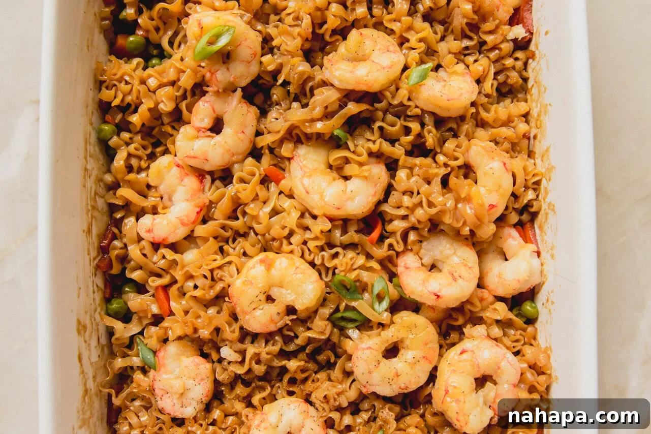 A white casserole dish with cooked shrimp on top of a ramen and vegetable sauce mixture.