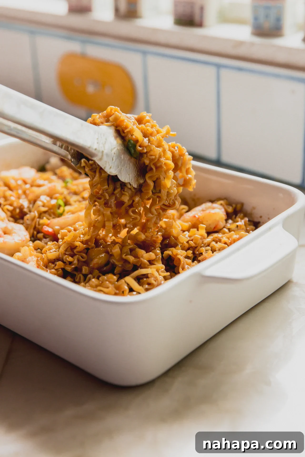 A pair of metal thongs lifting some of the shrimp ramen noodle bake out of the white ceramic dish it was baked it.
