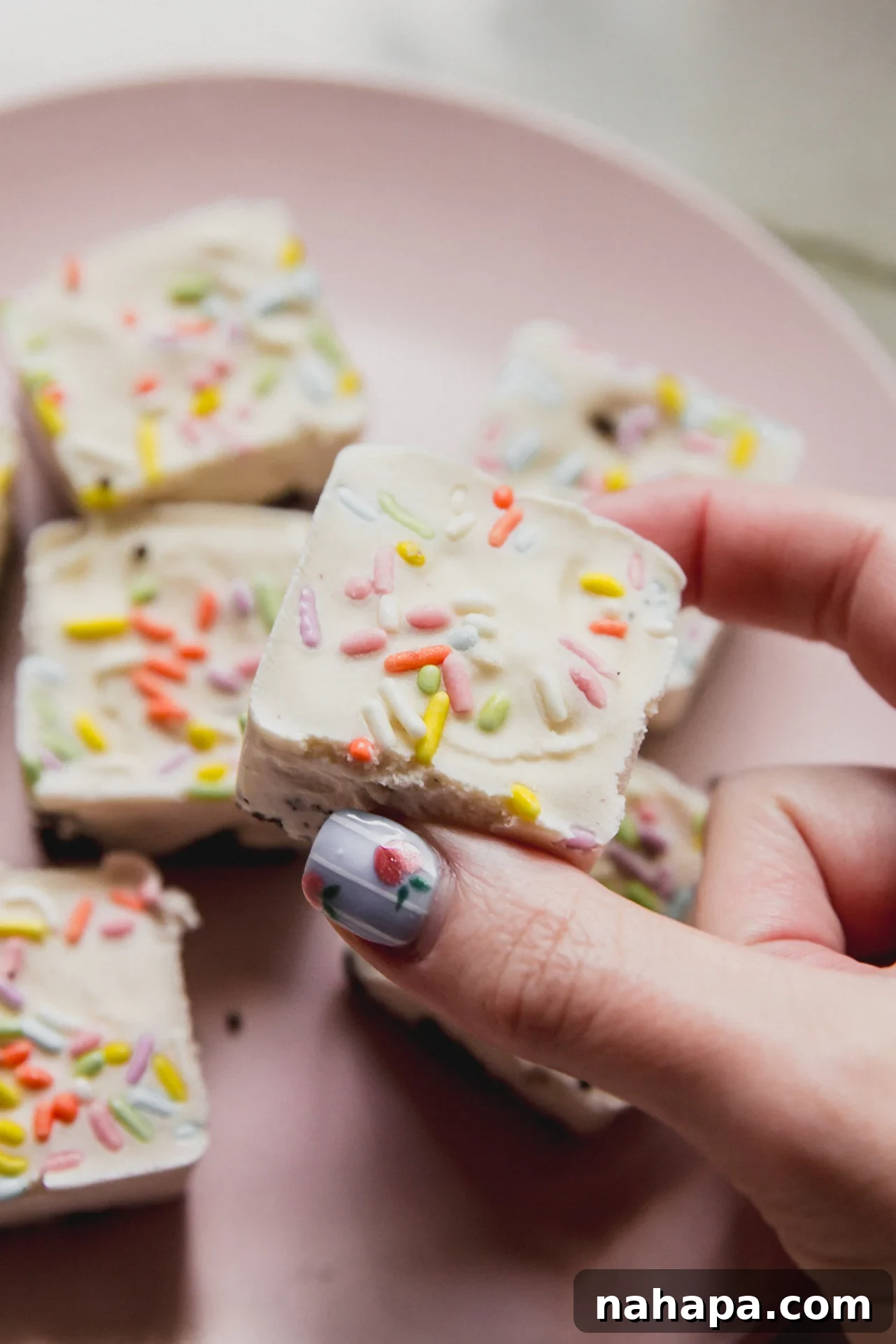 A hand holding a ice cream cake bite above a plate stacked with them.