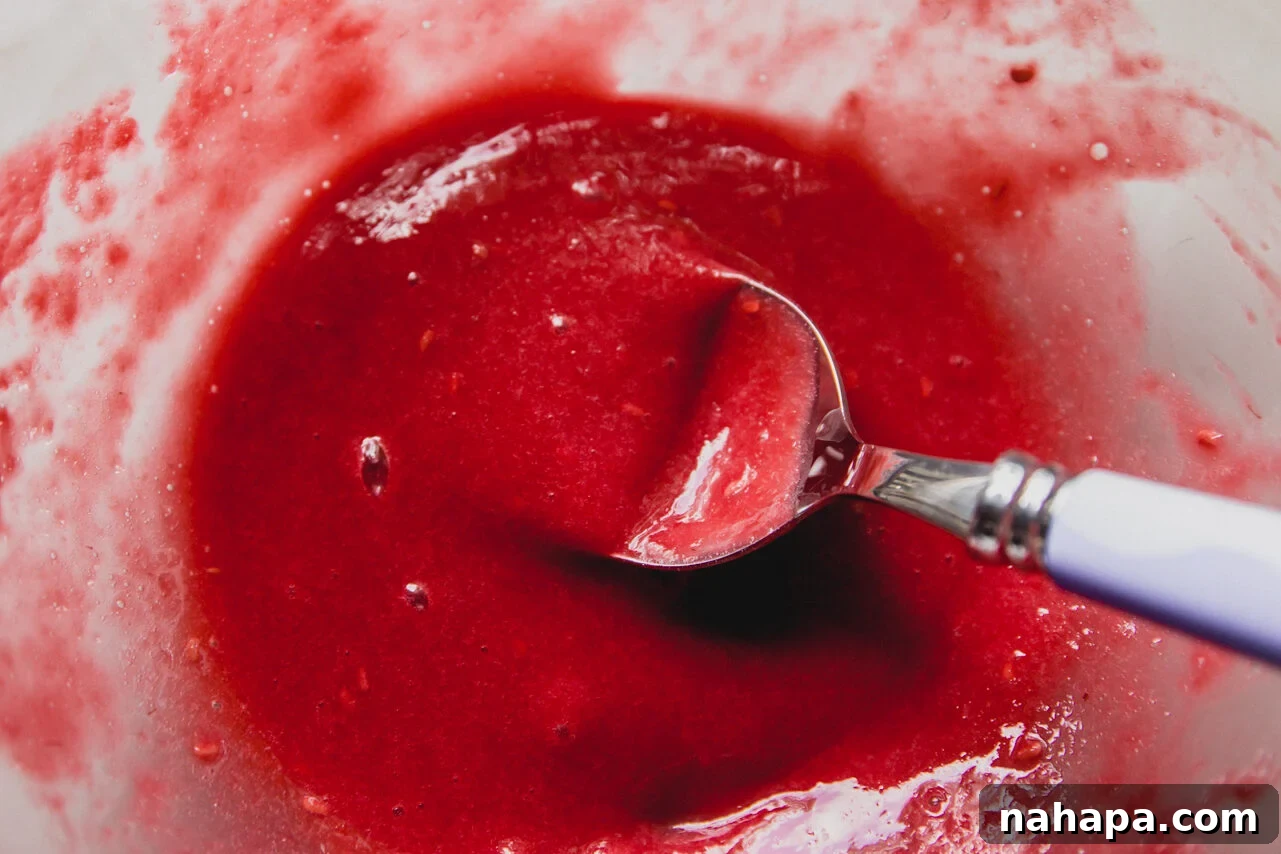 A glass bowl with raspberry mash being mixed by a spoon.
