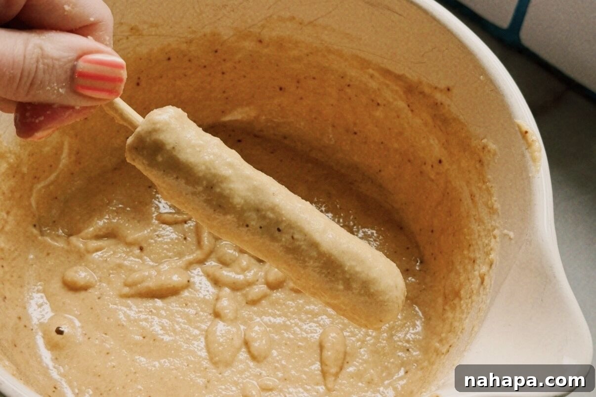 Gluten-Free Pancake and Sausage Sticks Recipe 6 A white ceramic bowl filled with pancake batter with a hand dipping one chicken sausage skewer into it to coat with batter.