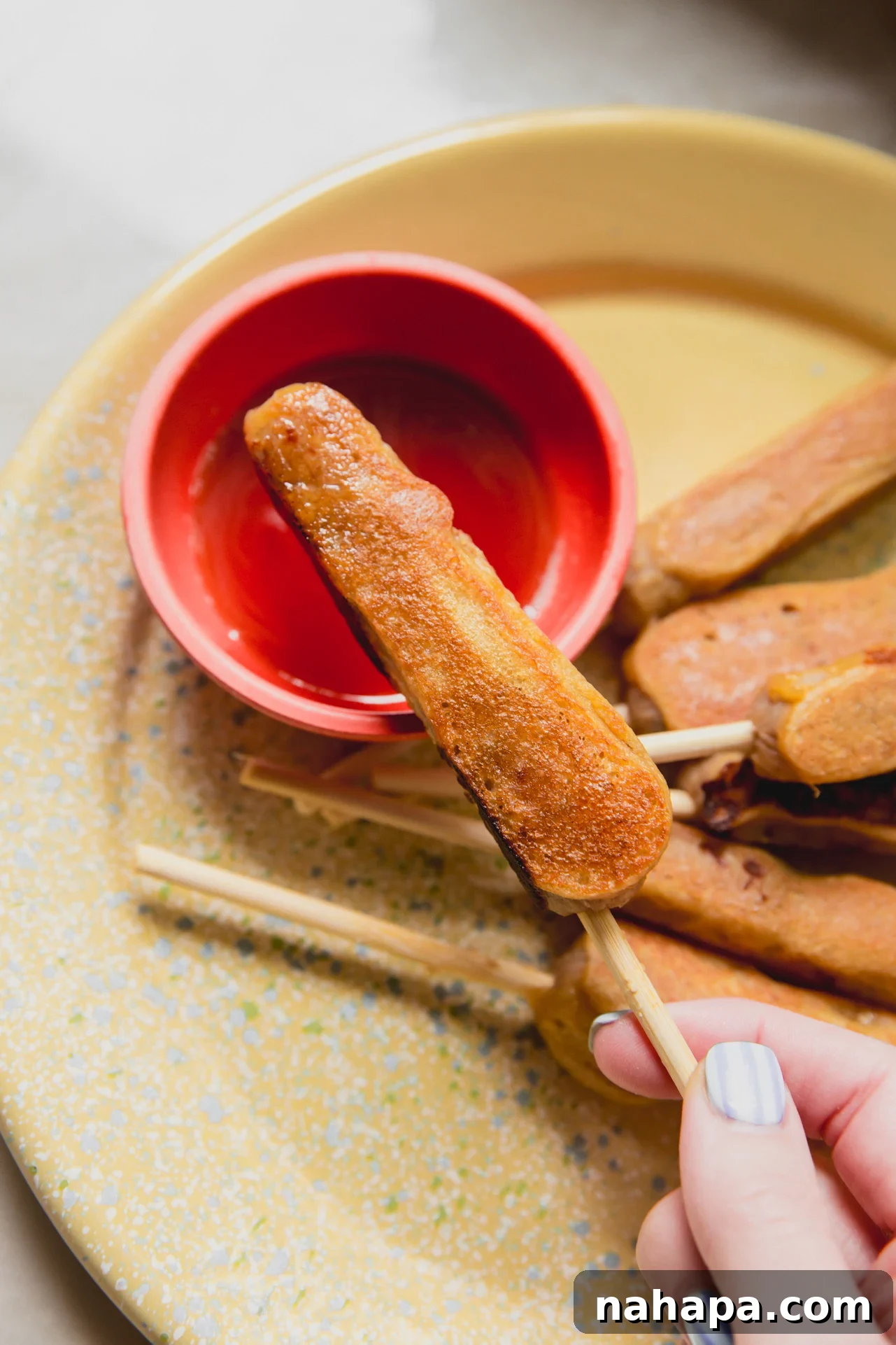Gluten-Free Pancake and Sausage Sticks Recipe 9 A hand holding a gluten-free pancake & sausage stick over a red bowl with maple syrup in it.