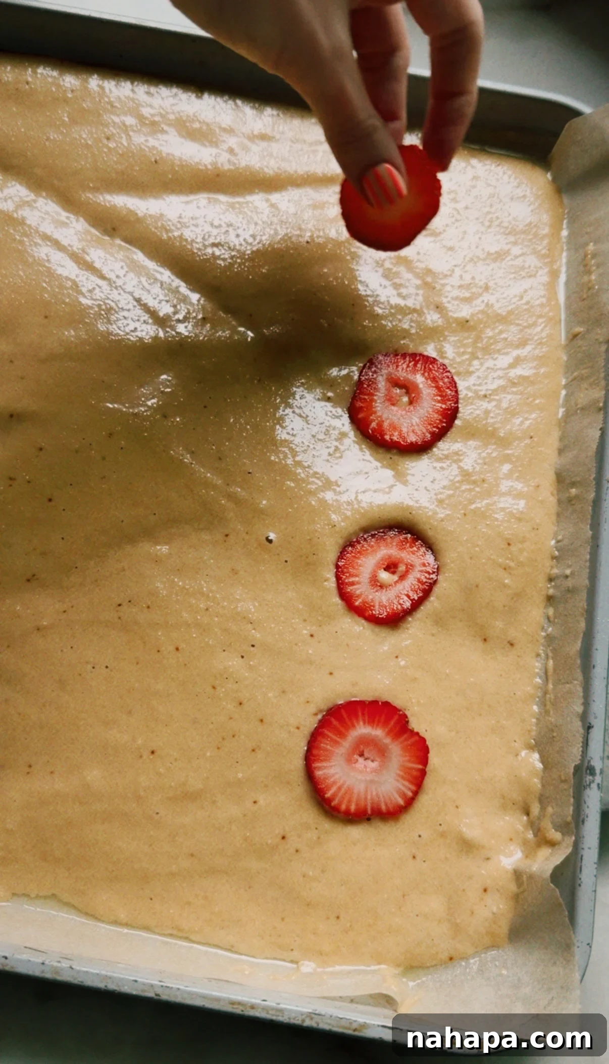 A hand placing sliced strawberries on unbaked sheet pan pancake batter
