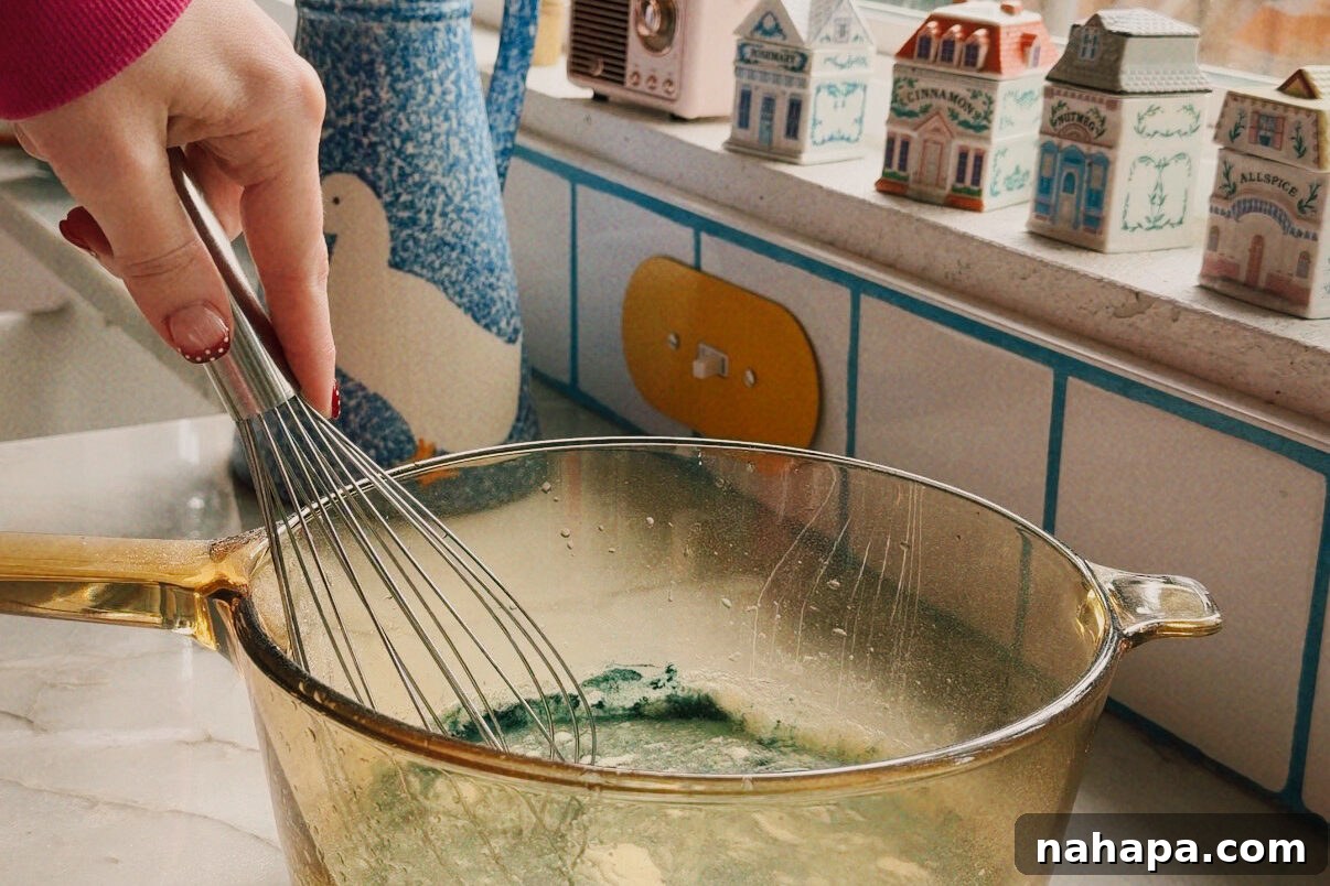 A glass bowl with apple juice, blue spirulina, and gelatin being mixed with a whisk.